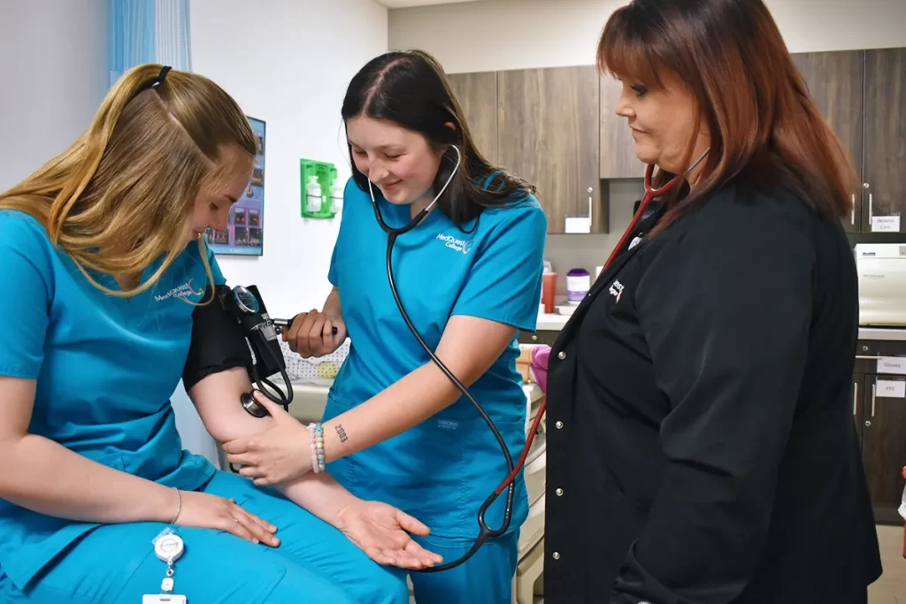medical assistant student taking blood pressure from another student while the teacher supervises